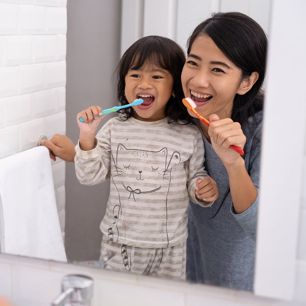 A woman and a child are brushing their teeth together in front of a bathroom mirror, smiling and enjoying their routine