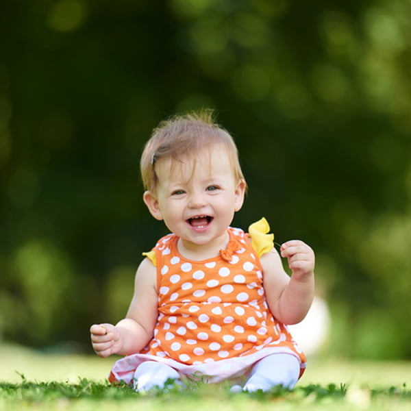 A baby girl in an orange dress sits on the grass, smiling and enjoying a sunny day outdoors