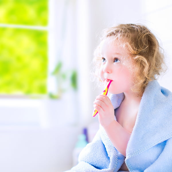 A young girl in a bathrobe brushes her teeth, smiling in a bright bathroom setting