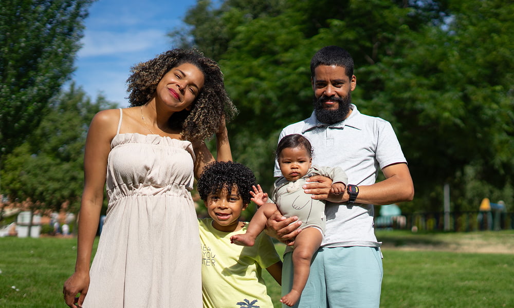 A family smiles together for a photo in a sunny park, surrounded by green trees and grass