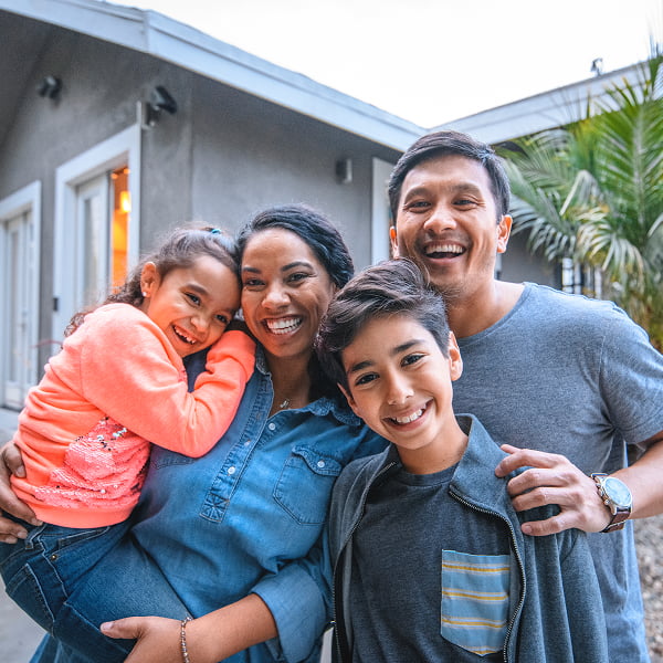 A family of four stands together in front of their home, smiling and posing for the camera