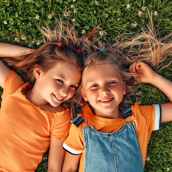 Two young girls lying on the grass, smiling joyfully at the camera under a clear blue sky