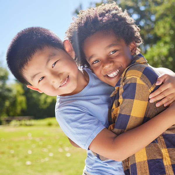 Two boys embrace each other warmly in a sunny park, surrounded by green grass and trees