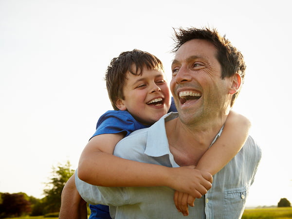 A father and son share a joyful laugh together in a sunny park, surrounded by green grass and trees