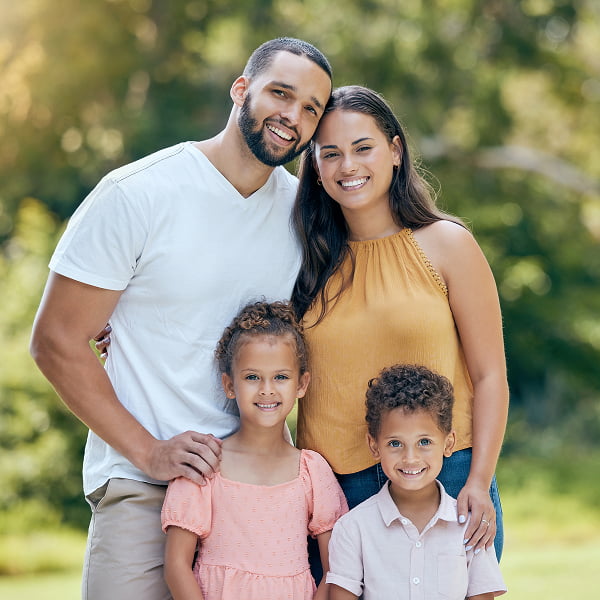 A family of four smiles for a photo in a sunny park, surrounded by green grass and trees