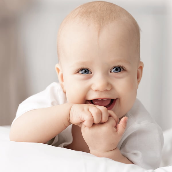A smiling baby lies on a bed, surrounded by soft bedding and a warm, inviting atmosphere