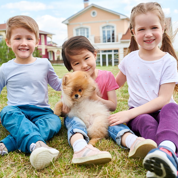 Three children sit on the grass, smiling and playing with a friendly dog beside them