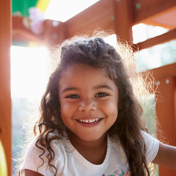 A young girl smiles while sitting on a wooden play structure, enjoying her time outdoors